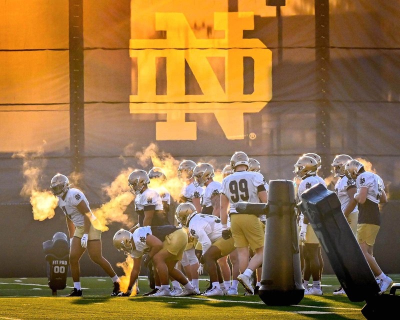 Football players train on a cold morning. The players breath can be seen in the air as a golden glow shines behind them.