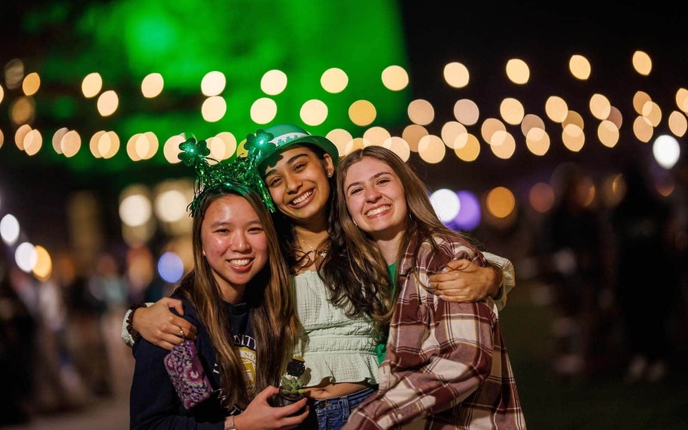 Students wearing St. Patrick's Day hats and headbands pose for a photo in front of blurred string lights.