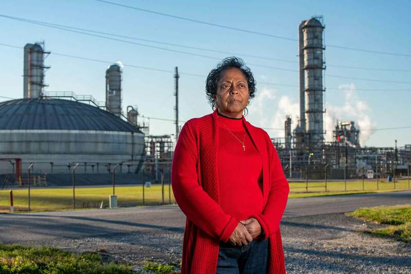 A woman wearing a red sweater and cross necklace stands outside in front of a chemical plant.