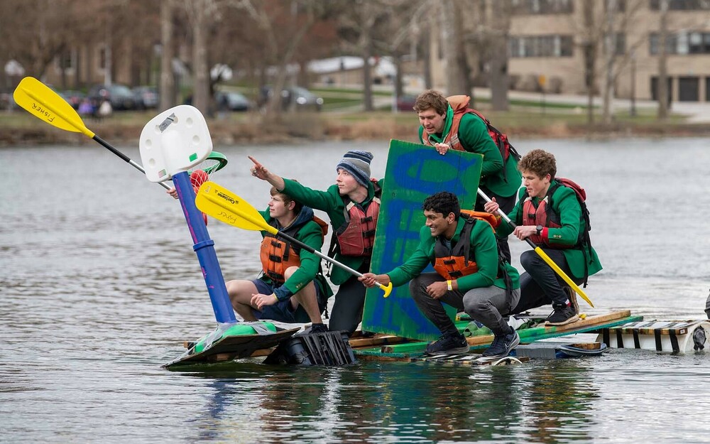 A group of students float on a makeshift boat made out of plastic tubs, wooden boards, and other various objects.