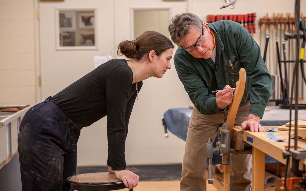 A professor shows a student carving details on a piece of furniture she's working on.