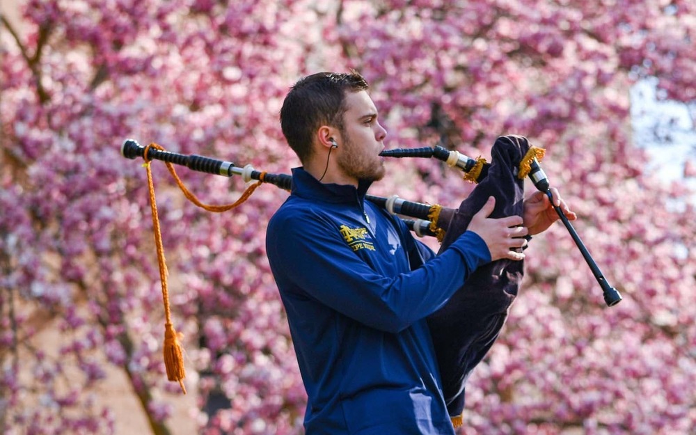 A student stands in front of a sea of pink flowers and plays bagpipes.
