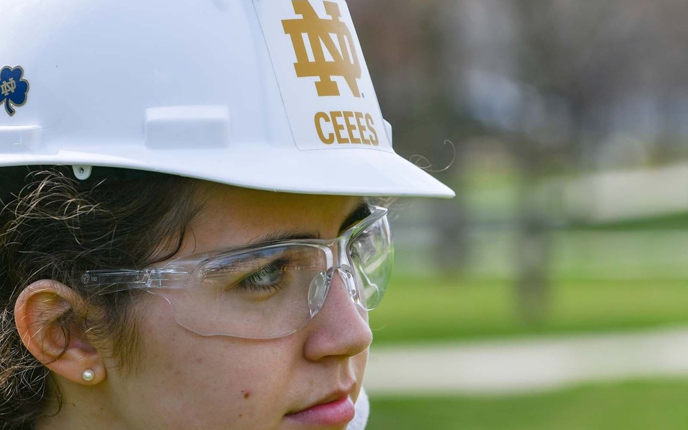 A student wears a hardhat with a Notre Dame CEES logo on it and safety glasses.