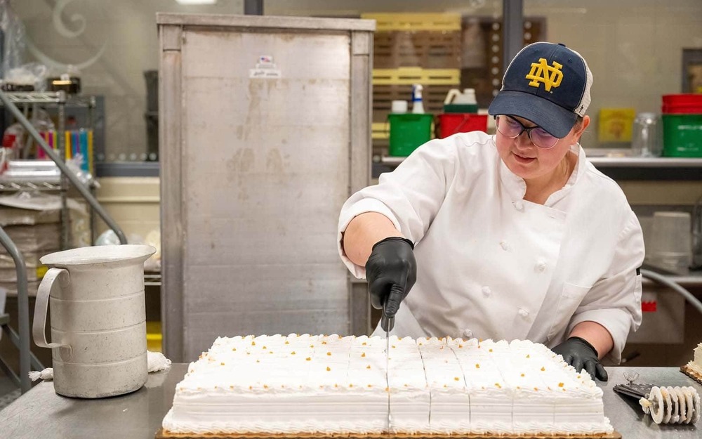 A white woman wearing a Notre Dame monogram hat cats a cake using a large knife.