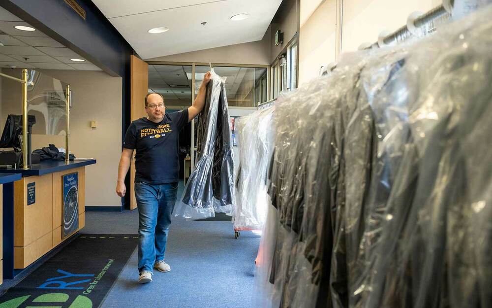A man wearing a Notre Dame Football t-shirt walks two graduation robes covered in plastic to a closet with other robes.