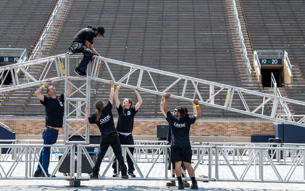 Six men assemble sections of stage made out of steel in the middle of a stadium. Five of the men hold up a section while another sits on top.
