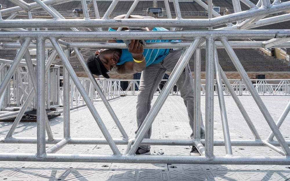 A black man bends over to tighten a bolt on a section of stage.