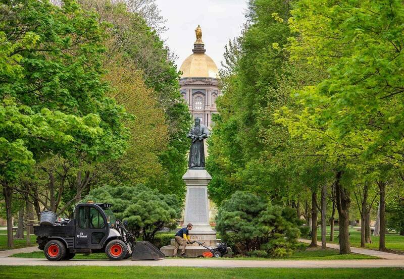 A man uses a tiller on soil in front of the Fr. Sorin Statue on the Main Quad. The Golden Dome is in the background surrounded by green trees.