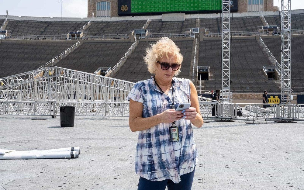 A white woman stands in the middle of the Notre Dame stadium,  in front of a stage being put together and checks her phone.