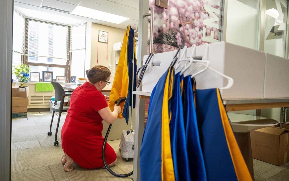 A woman wearing a red dress, kneels down and presses blue and gold honorary hoods in her office.