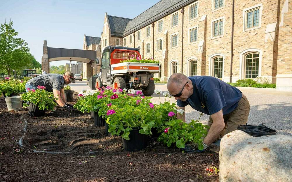 Two men plant flowers in a flower bed in front of the Morris Inn building.