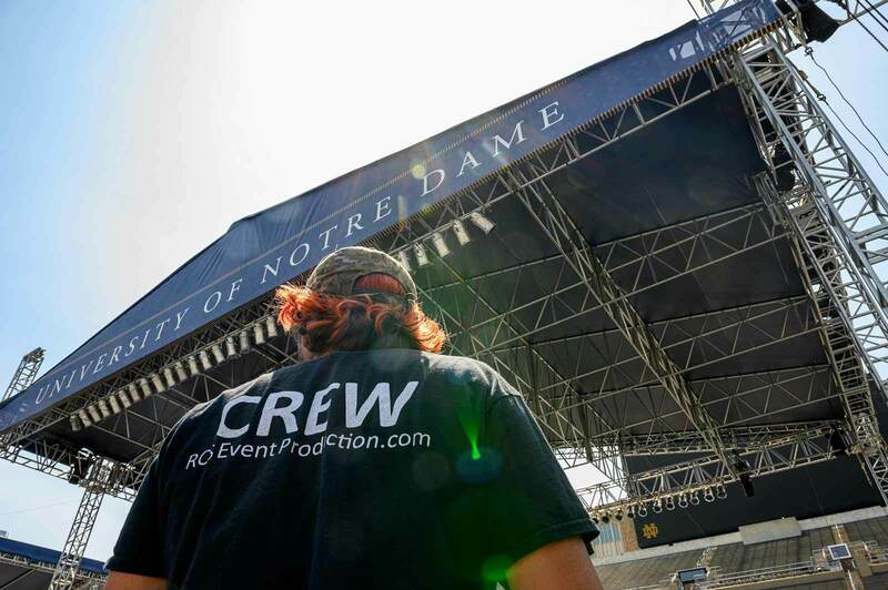 A photo of an individual from behind wearing a CREW shirt. In the background is a stage set up with a large University of Notre Dame banner on front.