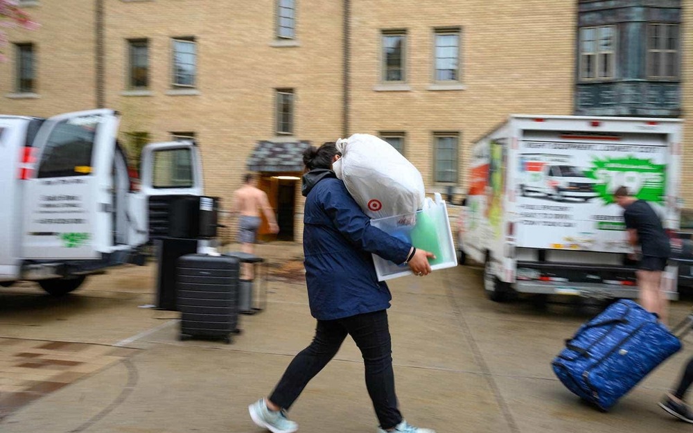 There are two moving vans and tubs in the background as a student walks past carrying a box and trash bag full of personal items.