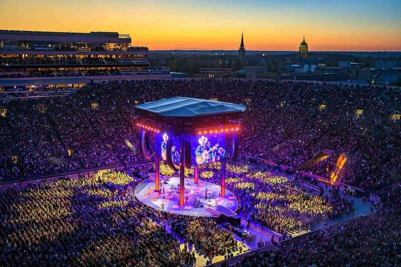 During sunset, Garth Brooks performs in Notre Dame Stadium. The stadium is dark besides the video board and colorful lights that shine on and around the stage. The Golden Dome and the Basilica can be seen off in the distance against the sunset.