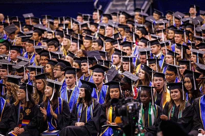 A large group of graduates wearing caps and gowns sit in the Purcell Pavilion at the Joyce Center.