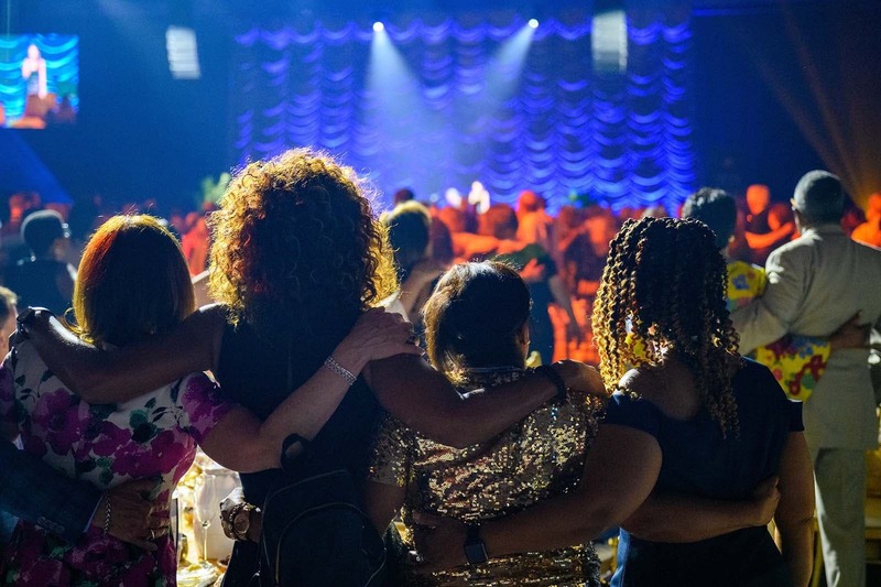 From behind, four women wrap their arms around each others shoulders and look towards a stage.