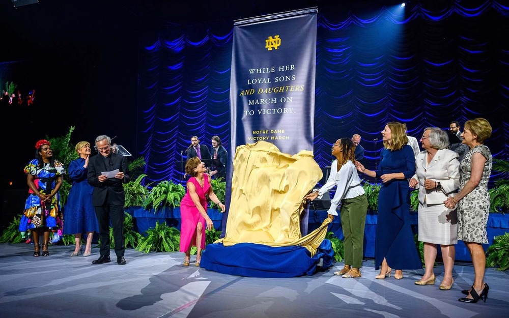 Two women unveiled a banner with new lyrics to the Notre Dame Victory March at the conclusion of the ‘Golden Is Thy Fame’ celebration gala dinner. Others around them cheer and clap.