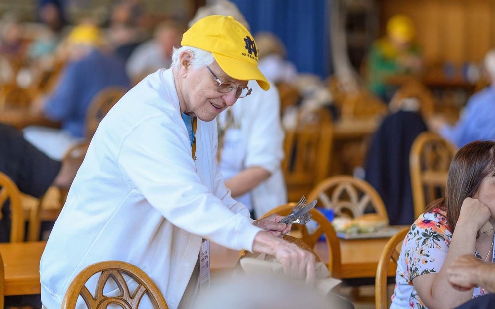 An elderly woman wearing a bright yellow Notre Dame hat places napkins and cutlery on a table.