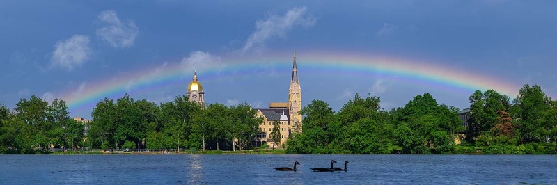 A rainbow stretches over St. Mary’s Lake, Notre Dame's Main Building and Basilica. Three geese are in the lake.