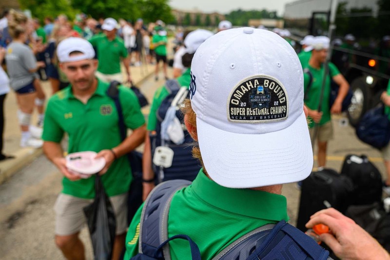 A close up of a baseball player wearing a College World Series hat backwards that says 'Omaha Bound, Super Regional Champs, NCAA Baseball.' In the background friends and family greet the Notre Dame Baseball team as they returned to campus after winning the NCAA super regional.