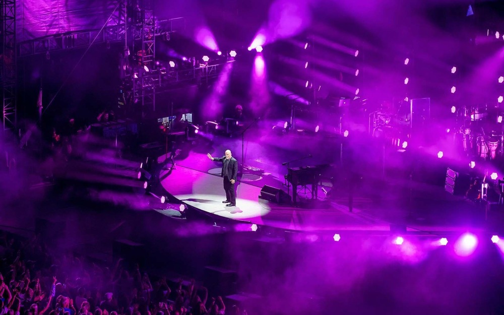 Billy Joel, on a glowing purple stadium, greets the crowd.