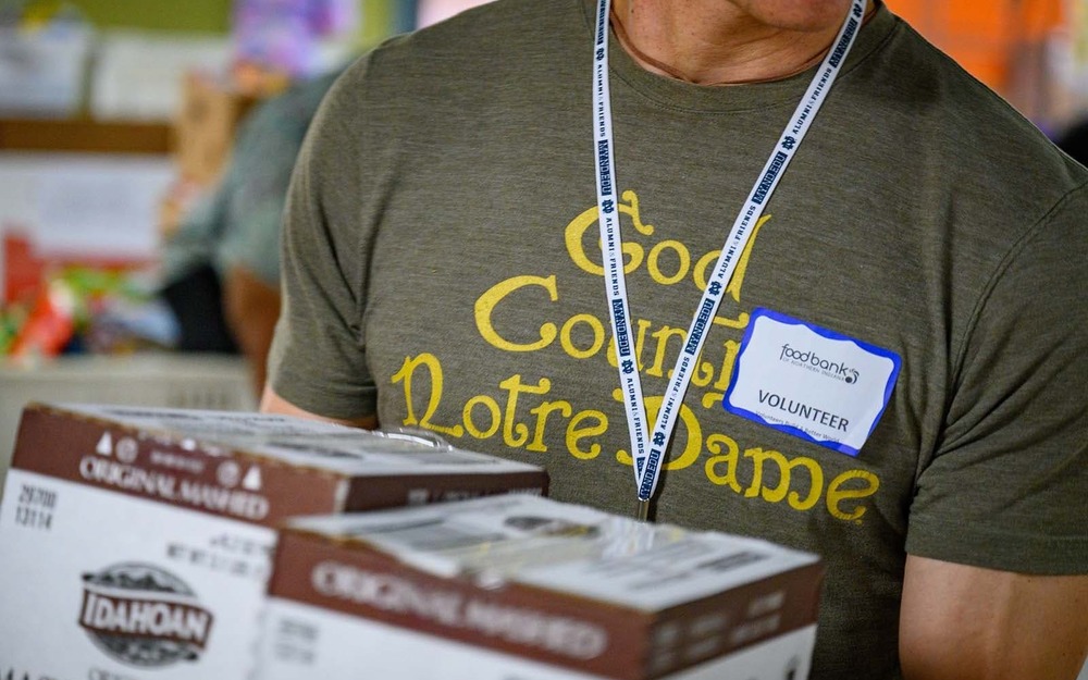 A close up of a man wearing name tag, lanyard, and a dark green shirt that says 'God, Country, Notre Dame' carries boxes.