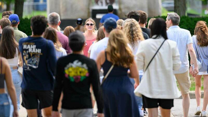 A woman wearing sunglasses walks backwards while talking and guiding a tour group outside.