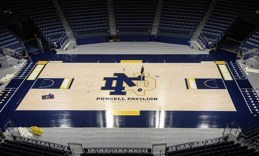 An individual works on painting the Notre Dame monogram on the floor of an empty basketball court.