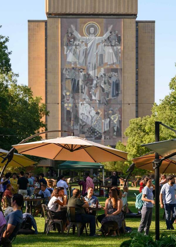 People sit at tables under large umbrellas on Library Lawn. In the background is The Word of Life Mural (Touchdown Jesus).