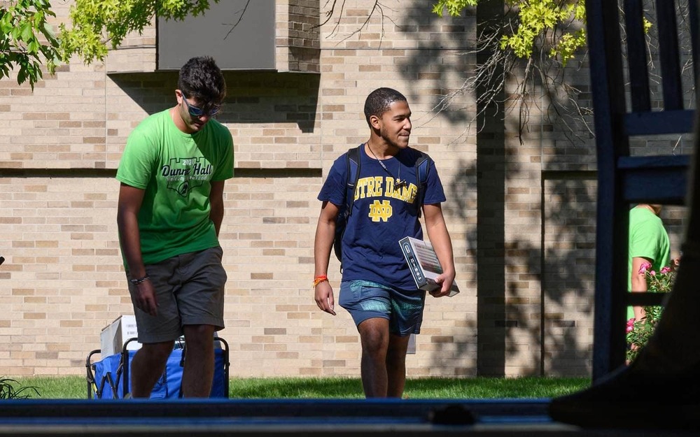 Two men wearing Notre Dame t-shirts, one carrying books to his side and the other pulling a cart walk on campus.
