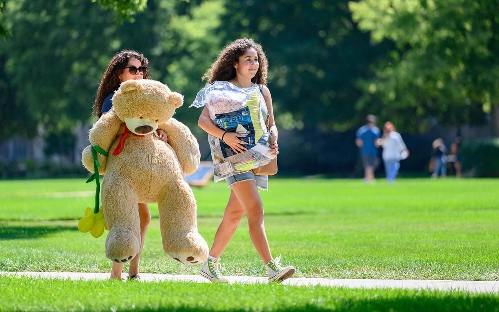 Two women walk carrying items for their dorm. One girl carries a large teddy bear.