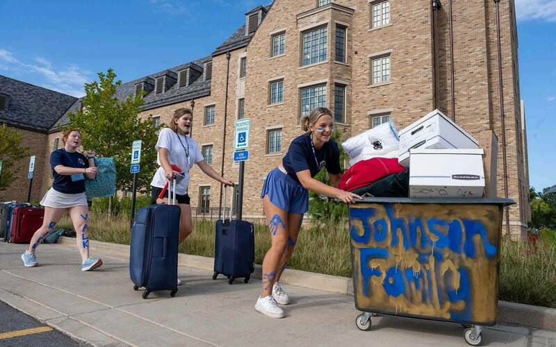 Three female students help with move-in. One student carries a bag, another pulls two suitcases, while the other pushes a large laundry cart full of items.