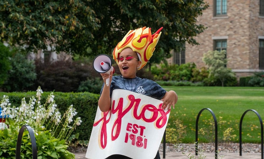 A girl yells into a megaphone while wearing a hat in the shape of fire and holding onto a 'Pyros Est. 1981' poster.