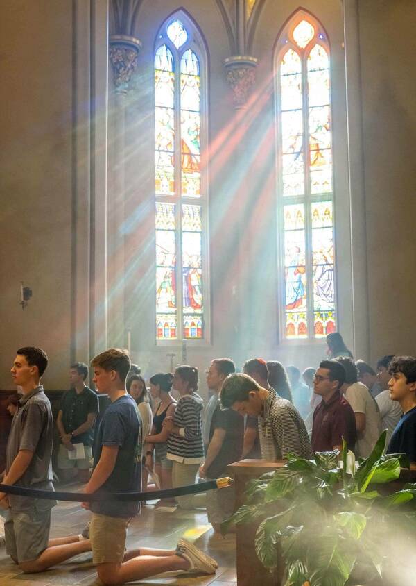 Students kneel inside of the Basilica of the Sacred Heart. Behind them sun rays shine in through stained glass windows.