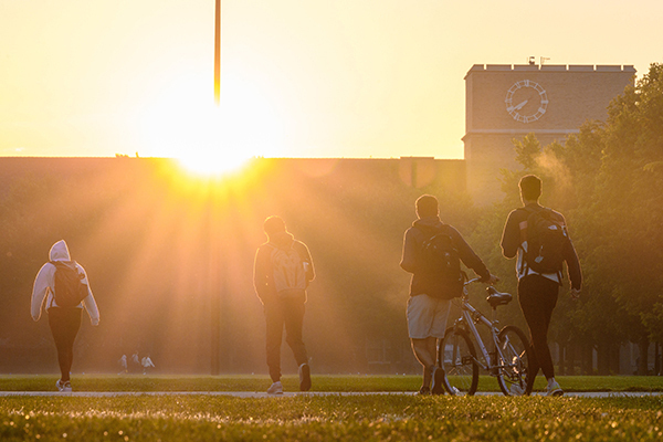 Four students walk across campus with the sun setting behind them.