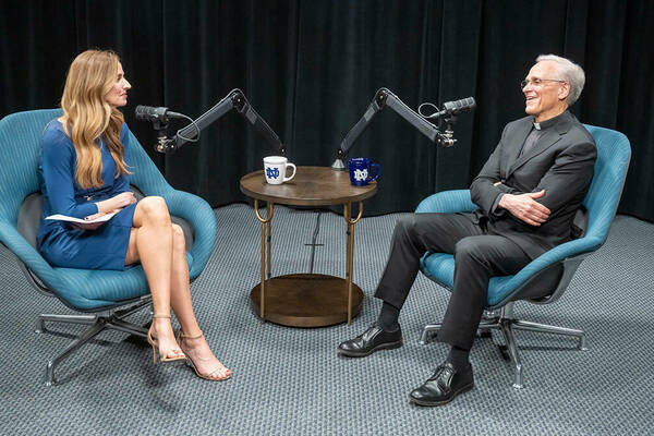 A blonde female in a blue dress sits smiling across from a smiling man with grey hair wearing a dark suit and a priest's collar. They are separated by a small circular table and two microphones. On the table are two Notre Dame coffee mugs: one white, one blue. Behind them is a black curtain backdrop.