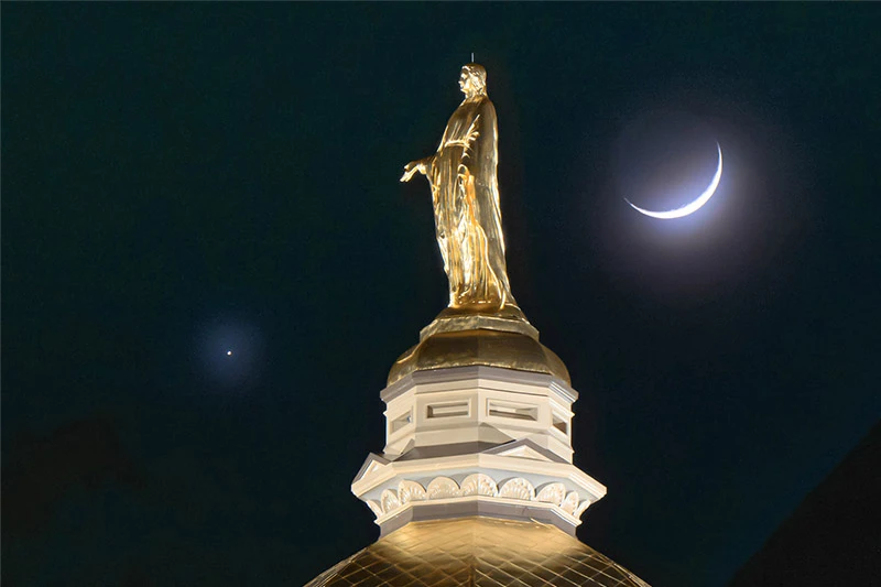 Golden statue of Mary with an outstretched arm atop Notre Dame's Main Building dome under a crescent moon and star.