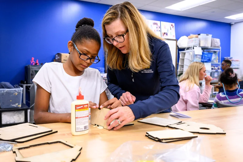 A woman in a  navy blue shirt helps a young girl wearing glasses assemble wooden craft pieces.