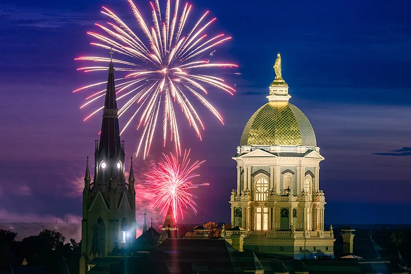 Fireworks burst over Notre Dame's Golden Dome and Basilica at dusk.