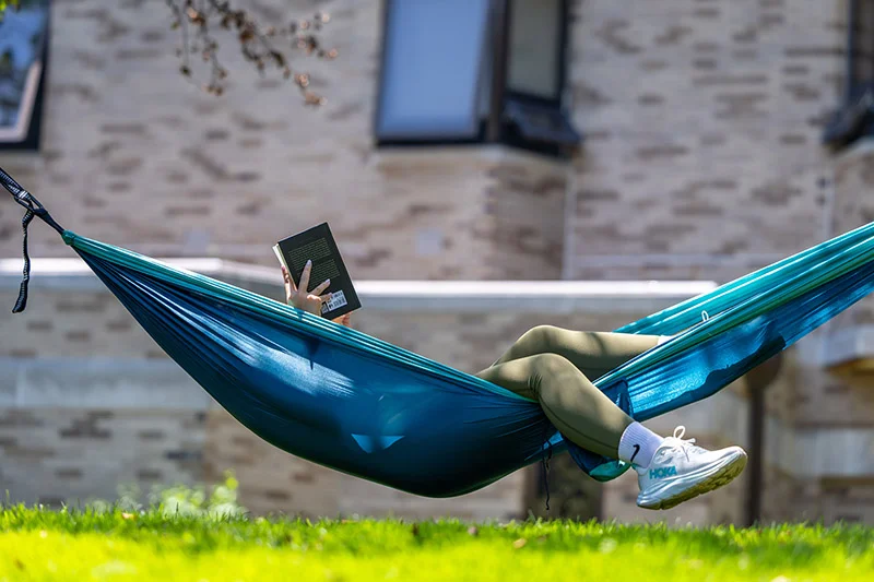 A person wearing green leggings and white sneakers lying in a teal hammock and reading a book on campus.