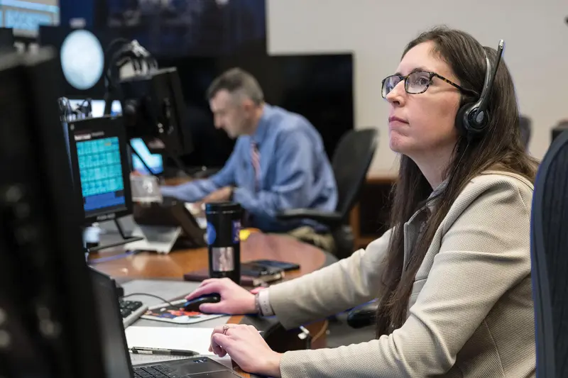 Focused woman in beige jacket, glasses, and headset, looking up while using a computer mouse. Man works at nearby station.