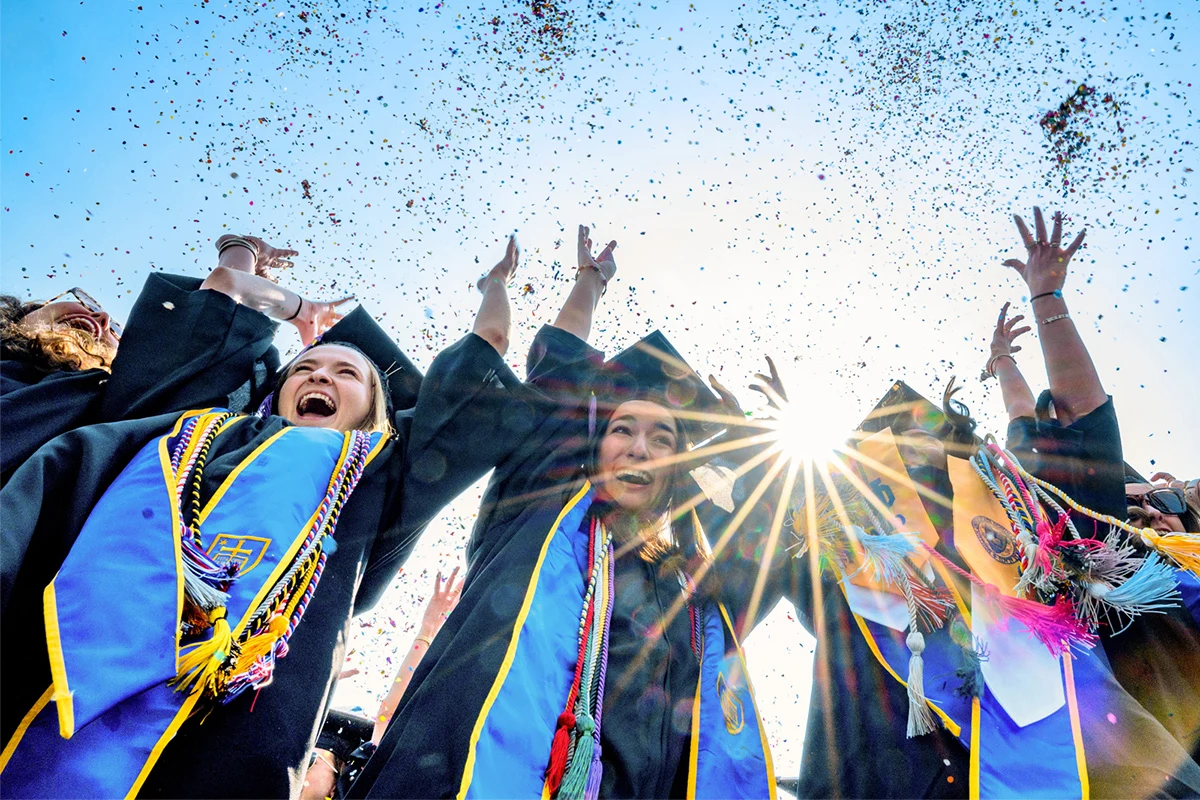 University of Notre Dame graduates in black caps and blue and gold regalia cheer with arms raised as colorful confetti fills the air during commencement ceremonies.