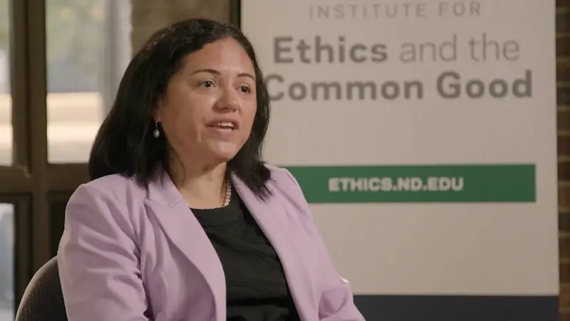 A woman in a lilac blazer speaks in front of a sign for the Institute for Ethics and the Common Good at Notre Dame.