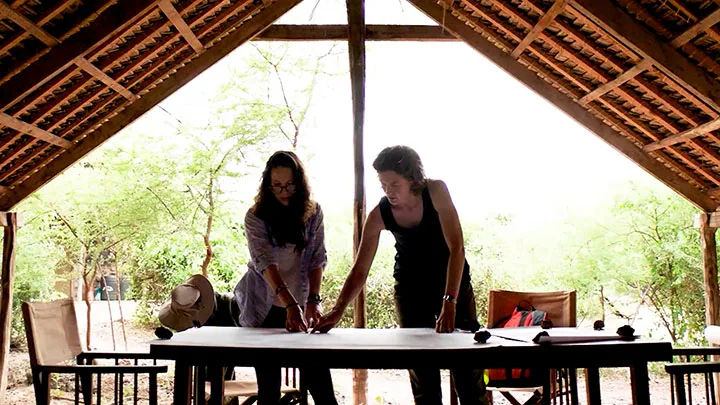 Researchers and archaeologists perform outdoor field work, unrolling maps under a thatched roof, surveying grassy landscapes, inspecting soil monitoring pipes, and delicately brushing away dirt from artifacts at an excavation site.