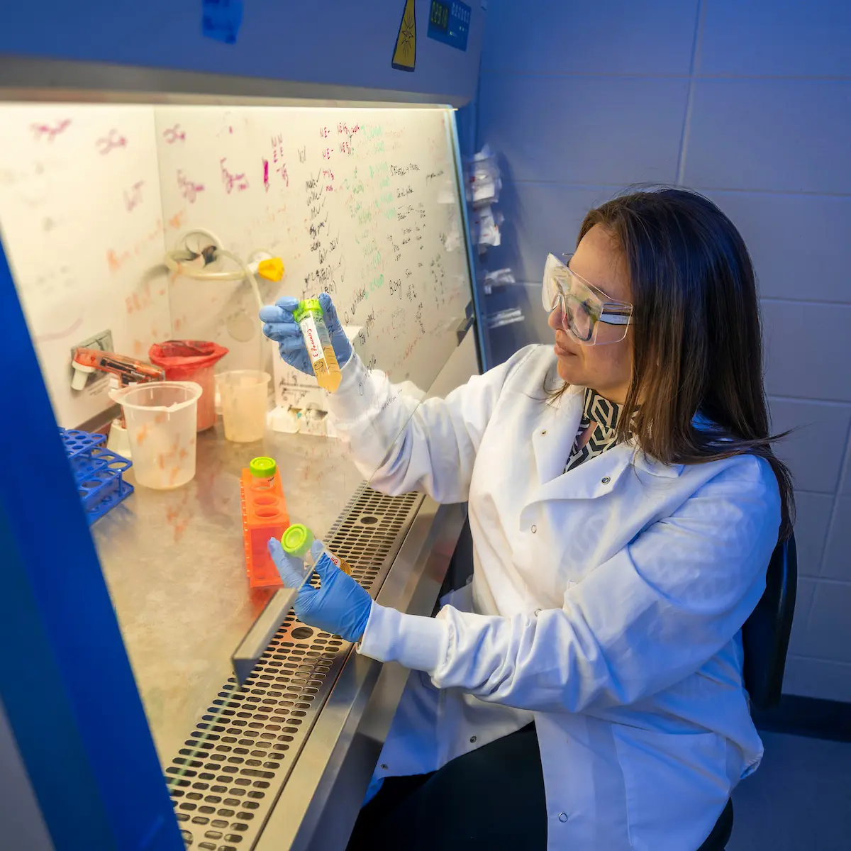 A woman in a white lab coat, blue gloves, and safety glasses holds up two vials, one with a yellow liquid and one with an orange liquid, in a lab.