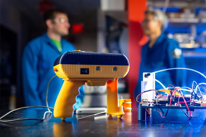 A yellow and navy electronic handheld scanner sits on a lab table next to exposed wiring and circuit boards, while two blurred lab workers in blue coats converse in the background.