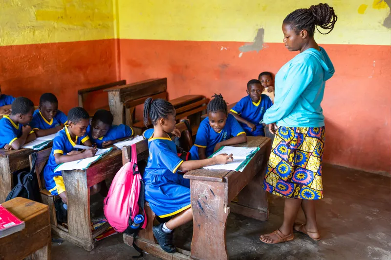 A teacher wearing a bright blue hoodie and patterned skirt stands over young students sitting at wooden desks in a classroom, helping them with their schoolwork.