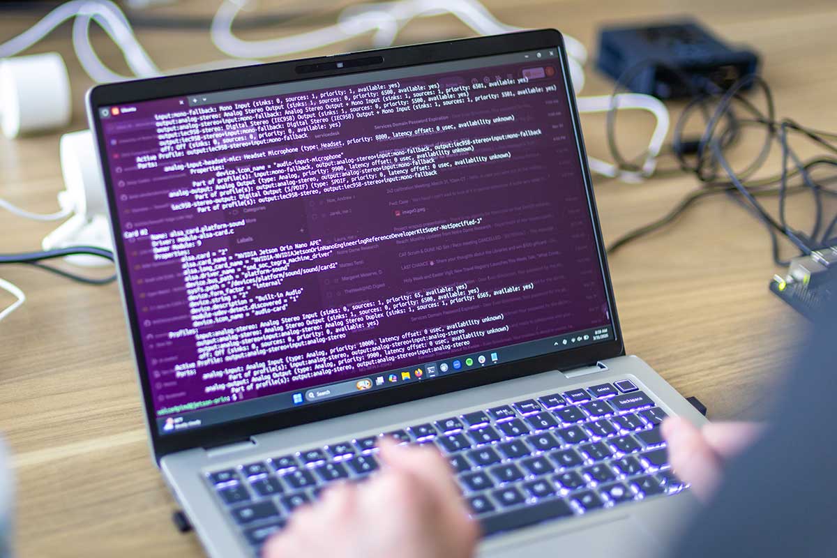 A close-up of hands typing on a laptop keyboard, with lines of code and system configuration text visible on the screen.