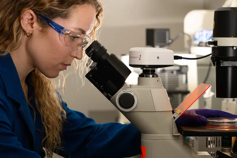 A young woman with long curly hair and safety goggles looks through a microscope in a lab, wearing a blue lab coat.