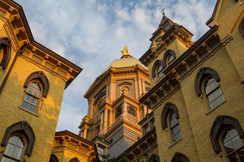 The golden dome of the Main Building at the University of Notre Dame, topped with a statue of Mary, rises above yellow brick buildings under a blue sky.